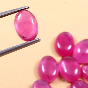 Puede incluir: Primer plano de una gema ovalada rosa sujeta con pinzas, con varias otras gemas del mismo color y forma dispersas en el fondo. Las gemas tienen una apariencia translúcida y una superficie pulida.