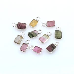 Puede incluir: Una colección de colgantes rectangulares de piedras preciosas en varios colores, incluyendo rosa, verde y amarillo. Cada colgante está engastado en un marco de metal plateado con un pequeño lazo en la parte superior para sujetarlo a una cadena. Las piedras tienen una textura natural.