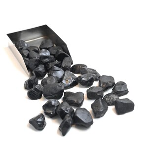 Puede incluir: Una colección de piedras preciosas negras rugosas que se derraman de una cuchara de acero inoxidable. Las piedras varían en tamaño y forma, con una superficie ligeramente reflectante. El fondo es blanco liso, resaltando el color oscuro de las piedras.