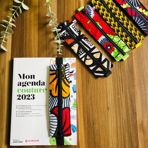 Peut inclure: Un livre noir et blanc intitulé "Mon agenda couture 2023" avec un marque-page en tissu coloré. Le marque-page a une fermeture éclair et un motif noir et blanc avec des accents rouges et jaunes. Le livre est sur une surface en bois avec plusieurs autres marque-pages en tissu coloré.