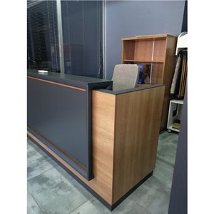 Puede incluir: Un mostrador de recepci&oacute;n moderno con una encimera gris oscura y un panel lateral de laminado de madera. El mostrador tiene un borde dorado.