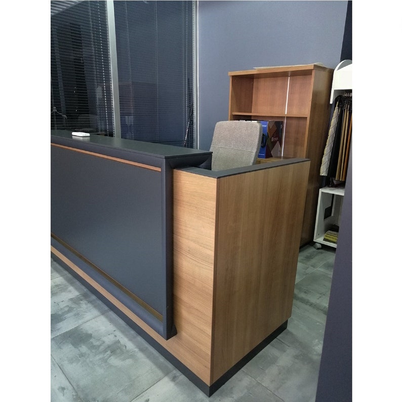 Puede incluir: Un mostrador de recepci&oacute;n moderno con una encimera gris oscura y un panel lateral de laminado de madera. El mostrador tiene un borde dorado.