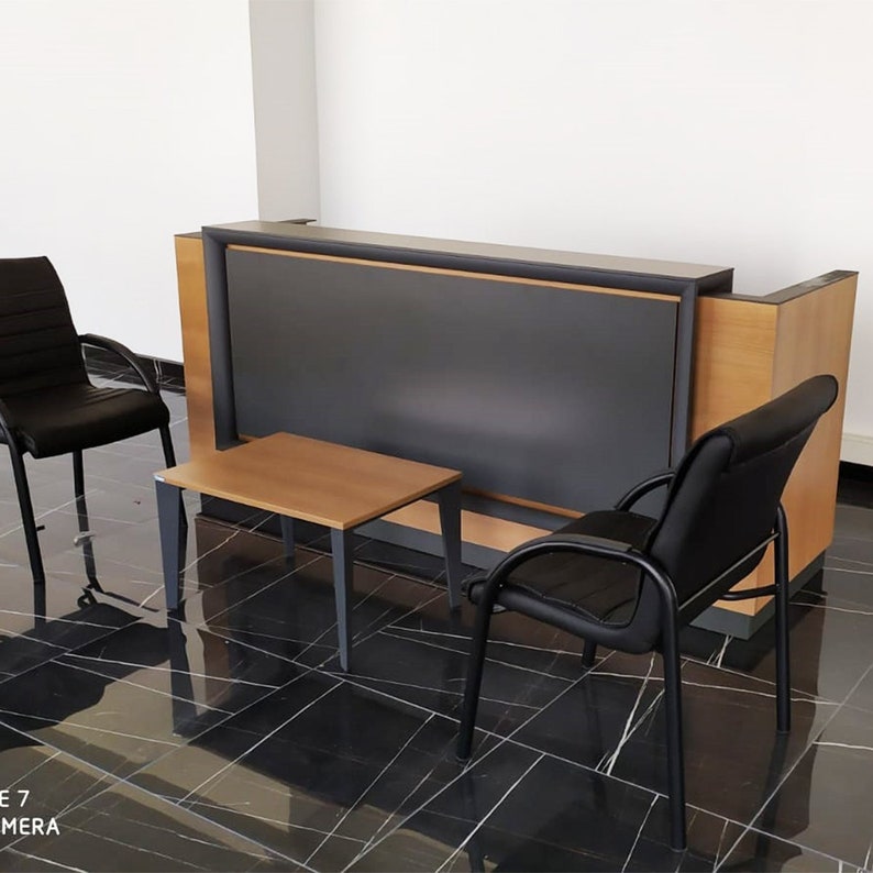 Puede incluir: Un mostrador de recepci&oacute;n negro con una encimera de laminado de madera y un panel frontal negro. Dos sillas negras con marcos de metal y una peque&ntilde;a mesa de madera con patas de metal negro est&aacute;n delante del mostrador.