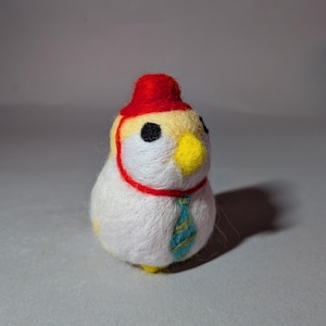 Op de afbeelding: Een naaldgevilte vogelbeeldje met een wit lichaam, gele kop en zwarte ogen. De vogel draagt een kleine rode hoed met een dunne rode kinband en een blauw met groen gestreepte das op de borst. De snavel is felgeel en de textuur is pluizig.