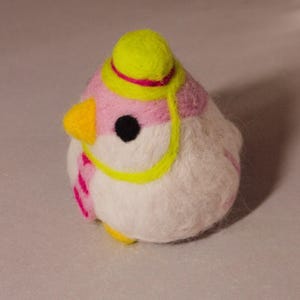 Op de afbeelding: Een naaldgevilte vogelbeeldje met een wit lichaam, roze accenten op de kop en een gele snavel. De vogel draagt een klein geel hoedje met een dunne roze band, een geel monocle-achtig detail over één zwart oog en een roze-witte gestreepte sjaal.