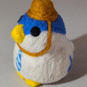 Op de afbeelding: Een naaldgevilte vogelbeeldje met een wit lichaam, blauwe kop en gele snavel. Het draagt een klein, rond, mosterdgeel hoedje met een dun riempje aan de zijkant. De vogel heeft zwarte ronde ogen en kleine gele pootjes.