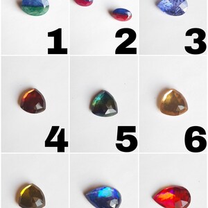May include: Nine different gemstone cabochons in various shapes and colors, including blue, green, red, brown, and gold. The cabochons are arranged in a grid pattern on a white background.