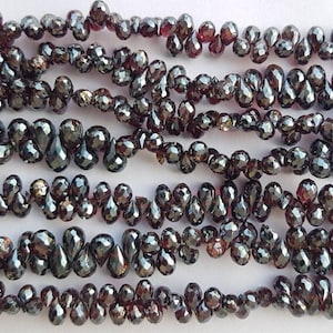 Puede incluir: Una hebra de cuentas de piedras preciosas de color rojo oscuro en forma de lágrima. Las cuentas son facetadas y tienen un acabado brillante y metálico.