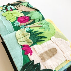 May include: A felt book with pages depicting a lush green jungle scene with palm trees, flowers, and a white building with a brown door. The book is handmade and features intricate details.