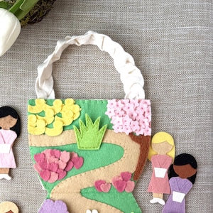 May include: Felt play set featuring a green felt background, a winding path, a pond, and a tree. There are felt figures of people and animals, including a girl in a pink dress, a girl in a purple dress, a boy in a yellow shirt, a boy in a blue shirt, and two white bunnies.