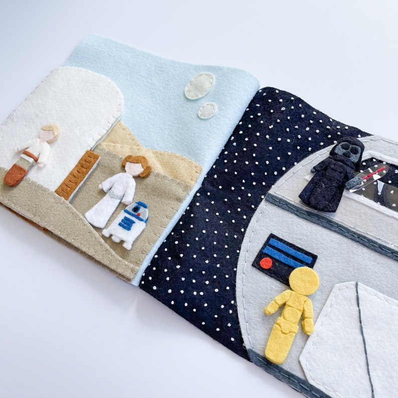 May include: A felt book with two pages. The left page shows a scene from Star Wars with Luke Skywalker, Princess Leia, and R2-D2. The right page shows a scene from Star Wars with Darth Vader, C-3PO, and the Millennium Falcon.