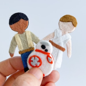 May include: Felt figures of Star Wars characters, including a brown-skinned human with a tan vest, a white-skinned human with a white robe, and a white droid with an orange stripe.