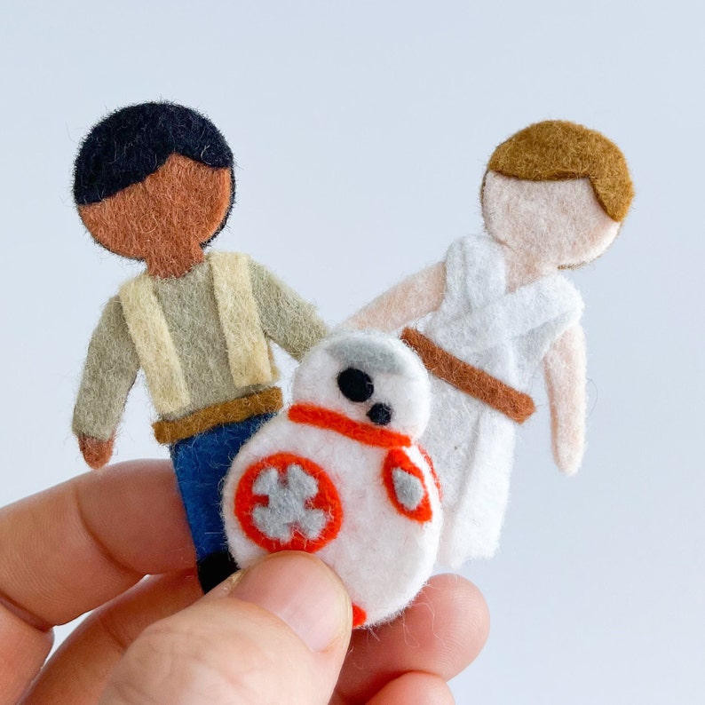 May include: Felt figures of Star Wars characters, including a brown-skinned human with a tan vest, a white-skinned human with a white robe, and a white droid with an orange stripe.