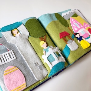 May include: A felt book with a princess theme. The book features a castle, a bridge, a well, and two princesses. The princesses are dressed in blue and yellow dresses. The book is made of felt and has a soft, plush texture.