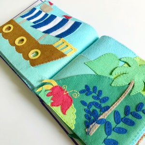 May include: A felt book with two pages. The left page features a brown sailboat with blue and white stripes on the sail. The right page features a green jungle scene with a palm tree, leaves, and a red flower.