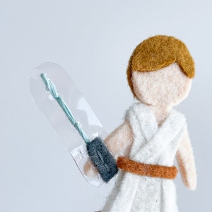 May include: A felt doll of a character from Star Wars, wearing a white robe with a brown belt and holding a lightsaber in a clear plastic case.