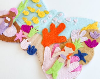 May include: A felt mermaid-themed play set with two mermaids, coral, seaweed, and fish. The set is made of felt and has a light blue background.