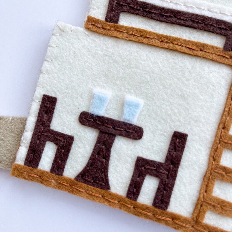 May include: A felt craft depicting a small table with two chairs and two glasses. The table and chairs are brown, and the glasses are light blue. The background is white.