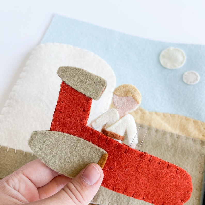 May include: A felt toy spaceship with a red and beige body and a beige cockpit. A felt toy character with blonde hair and a white shirt is sitting in the cockpit.