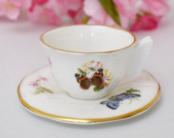 May include: A white teacup and saucer set with a gold trim. The teacup features a butterfly design with orange, black, and white wings. The saucer has a blue butterfly design.