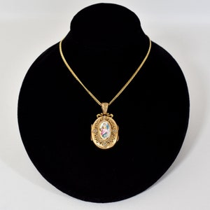 May include: A gold-colored oval locket necklace with a detailed design, featuring a miniature painting of a woman in a dress. The locket hangs from a delicate gold chain, displayed on a black velvet bust.