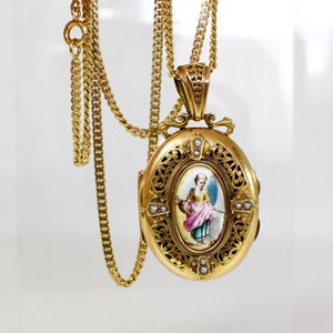 May include: A gold-toned oval locket pendant with a detailed filigree design, suspended from a matching chain. The locket features a miniature painting of a woman in a pink dress. Small pearls are set around the painting.