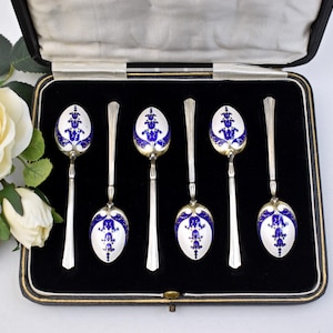 May include: Set of six antique silver spoons displayed in a black velvet-lined case. Each spoon features a white bowl with a blue floral design and a long, slender handle. The spoons are likely made of sterling silver.