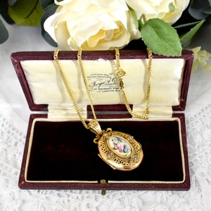 May include: A gold-toned oval locket necklace with a detailed pendant featuring a painted image and small pearls. The locket is displayed open inside a burgundy velvet-lined box. The necklace chain is delicate and gold.