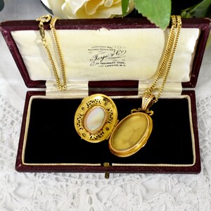 May include: Open gold locket necklace with a chain, displayed in a burgundy velvet box. The locket has an oval shape with a mother-of-pearl center and intricate filigree detailing. The box has the text "Toye & Co" printed on the inside.