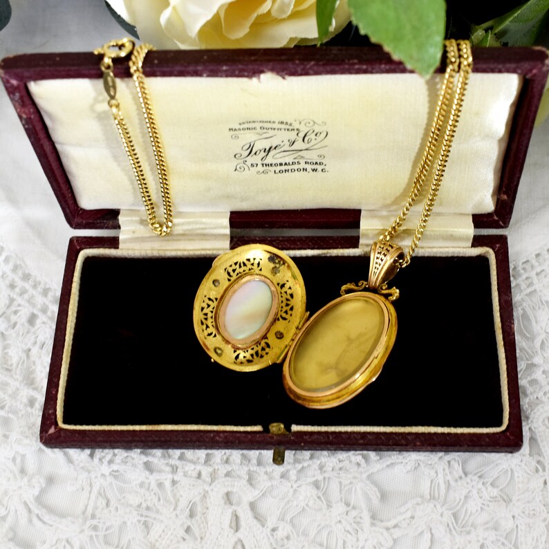 May include: Open gold locket necklace with a chain, displayed in a burgundy velvet box. The locket has an oval shape with a mother-of-pearl center and intricate filigree detailing. The box has the text "Toye & Co" printed on the inside.