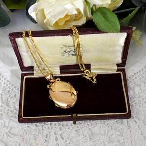 May include: A gold-colored oval locket pendant on a delicate chain, displayed open in a burgundy velvet-lined box. The locket has a decorative top and is set against a cream-colored interior. The box is open, revealing the necklace.