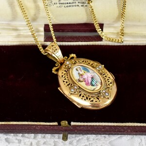 May include: A gold-toned oval locket pendant with a detailed filigree design and a small painted portrait. The pendant is suspended from a delicate gold chain. Small pearls are set around the portrait. The locket is displayed in a burgundy velvet box.