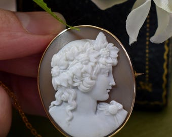 May include: Oval cameo pendant with a profile of a woman with a floral crown, set in a gold frame. The cameo is a light gray color, and the gold frame has a delicate chain attached. The background is a soft brown.