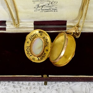 May include: Open gold locket pendant with a delicate chain. The locket features an oval mother-of-pearl center and intricate filigree detailing. The locket is displayed open, revealing its interior. The chain is a fine, gold curb link style.