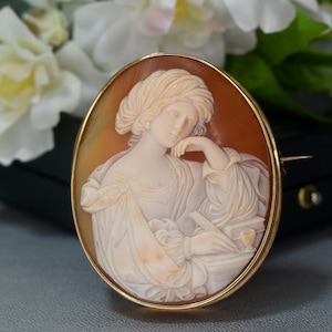 Peut inclure: Broche camée ovale avec un cadre doré, représentant un relief sculpté d'une femme. La femme est de profil, avec des cheveux et des vêtements détaillés, tenant un instrument d'écriture. Le fond est de couleur chaude et terreuse.