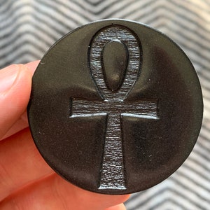 May include: A black circular coaster with a raised, textured ankh symbol in the center.