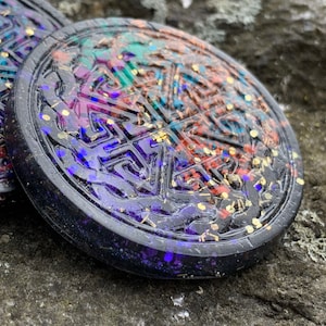 May include: Two round, black resin coasters with a Celtic knot design. The coasters are decorated with iridescent glitter and gold flakes. The design is a swirling pattern of blue, purple, green, and orange.