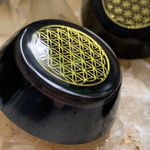 May include: A black obsidian crystal sphere with a gold flower of life design etched on the top. The sphere is sitting on a white crystal surface.