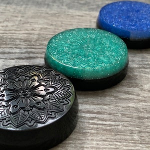 May include: Three round coasters with a black base and a decorative top. The first coaster has a black and silver mandala design. The second coaster has a teal green glitter top. The third coaster has a blue glitter top.