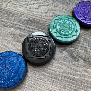 May include: Four round, decorative coasters with a geometric design in the center. The coasters are made of black, blue, green, and purple resin with a glittery finish.