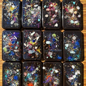 May include: Twelve small rectangular black resin tiles with embedded colourful glitter and small pieces of iridescent glass. The tiles are arranged in four rows of three.