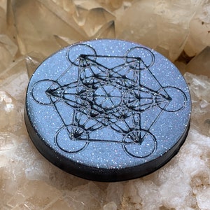 May include: A round, black, glitter-covered coaster with a silver metallic geometric design. The design is a sacred geometry symbol known as the Flower of Life.