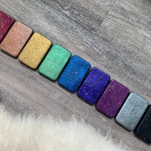 May include: A row of nine small rectangular boxes with different glitter colors, including burgundy, peach, gold, green, blue, purple, silver, and black.