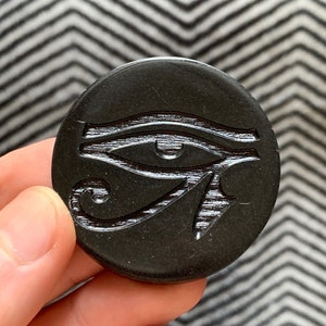 May include: A black circular pendant with a raised, detailed carving of the Eye of Horus, an ancient Egyptian symbol.