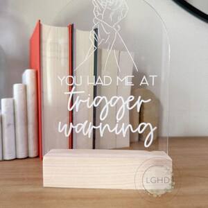 May include: Clear acrylic sign with the text "YOU HAD ME AT trigger warning" in white script. The sign features a line drawing of two hands clasped together. The sign is mounted on a natural wood base.