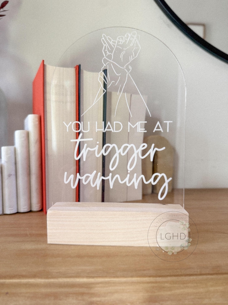 May include: Clear acrylic sign with the text "YOU HAD ME AT trigger warning" in white script. The sign features a line drawing of two hands clasped together. The sign is mounted on a natural wood base.
