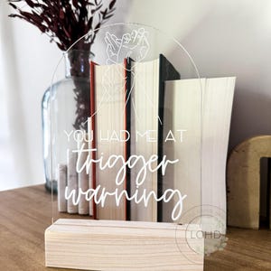 May include: Clear acrylic sign with the text "YOU HAD ME AT trigger warning" in white script. The sign features a hand gesture design and sits on a natural wood base. Books are visible behind the sign, and a vase with dried flowers is in the background.