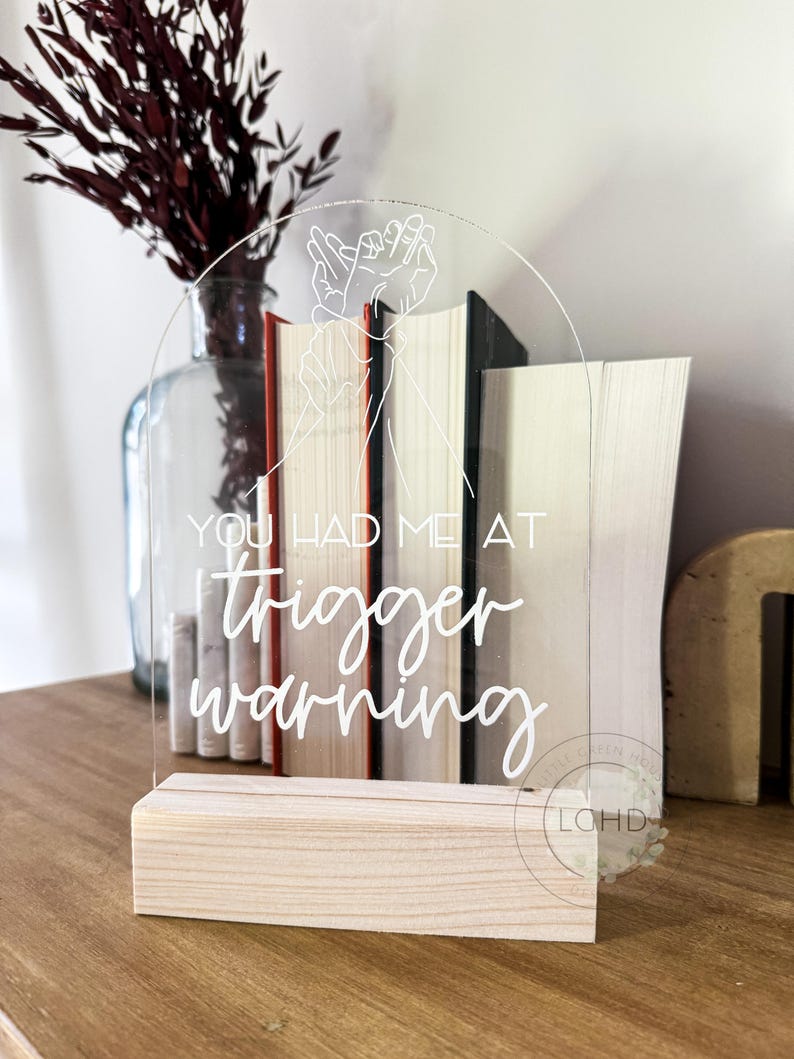 May include: Clear acrylic sign with the text "YOU HAD ME AT trigger warning" in white script. The sign features a hand gesture design and sits on a natural wood base. Books are visible behind the sign, and a vase with dried flowers is in the background.