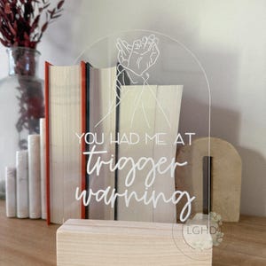 May include: Clear acrylic sign with the text "YOU HAD ME AT trigger warning" in white script. The sign features a line drawing of two hands clasped together. The sign is set in a light wood base.