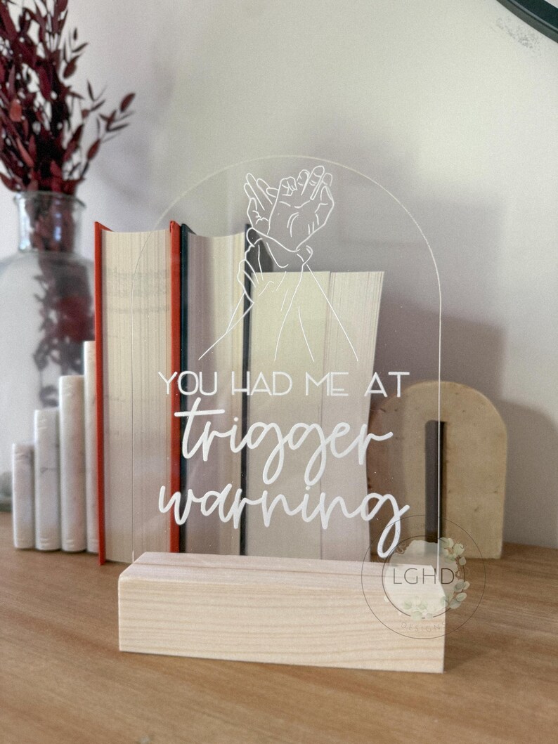 May include: Clear acrylic sign with the text "YOU HAD ME AT trigger warning" in white script. The sign features a line drawing of two hands clasped together. The sign is set in a light wood base.